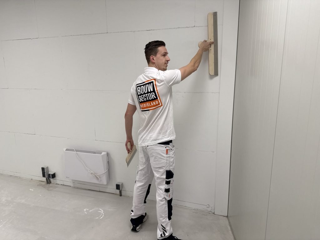 Stukadoor in wit poloshirt van Bouwsector Nederland werkt met spaan en plakspaan aan een lege kamer