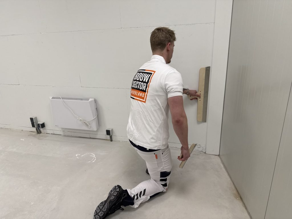 Stukadoor van Bouwsector Nederland zit op een knie en werkt de onderkant van een muur glad af