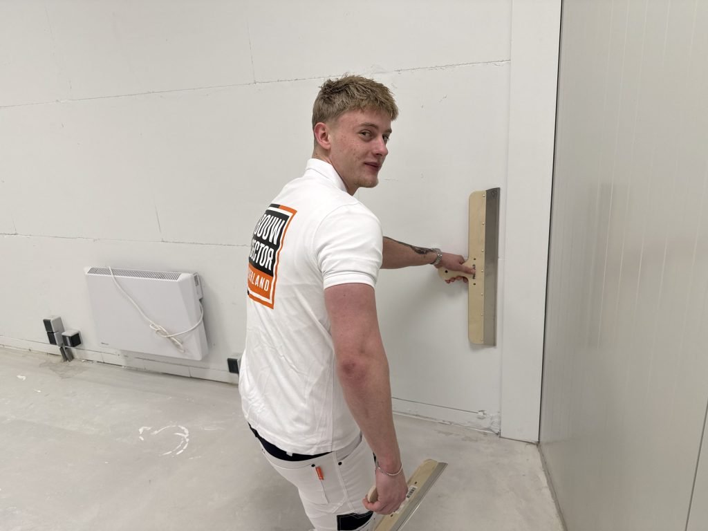 Stukadoor met Bouwsector-logo op zijn shirt zet een brede spaan tegen een kale binnenmuur